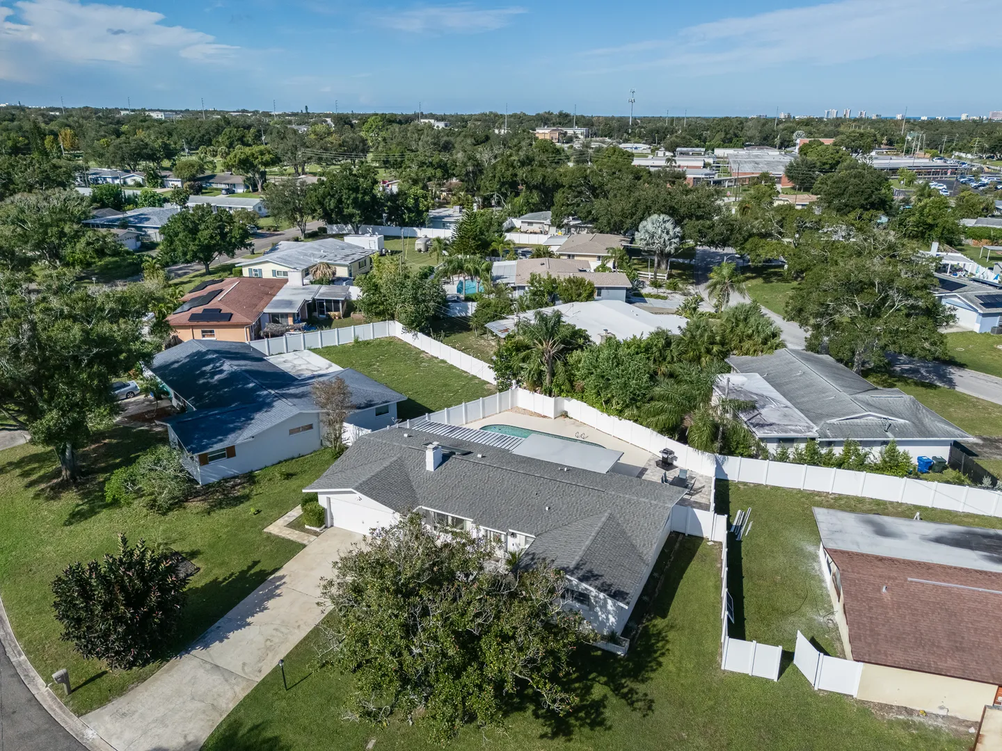 Aerial view of a single-story house with a fenced backyard, swimming pool, and driveway surrounded by a residential neighborhood.
