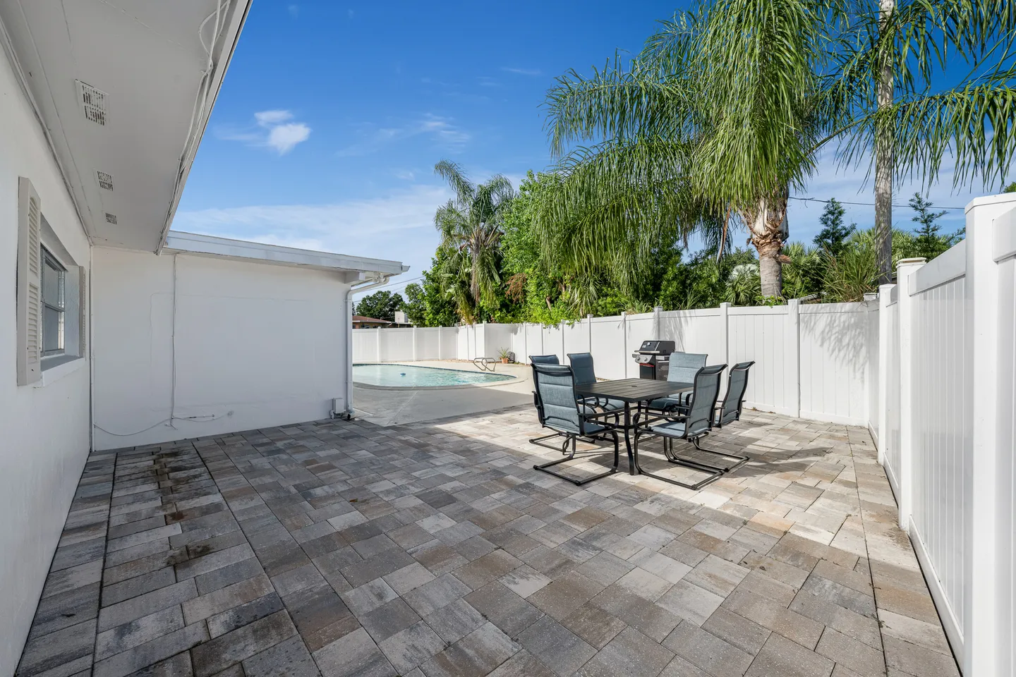 Outdoor paved patio area featuring a dining set and barbecue grill with a swimming pool in the background enclosed by a white fence.