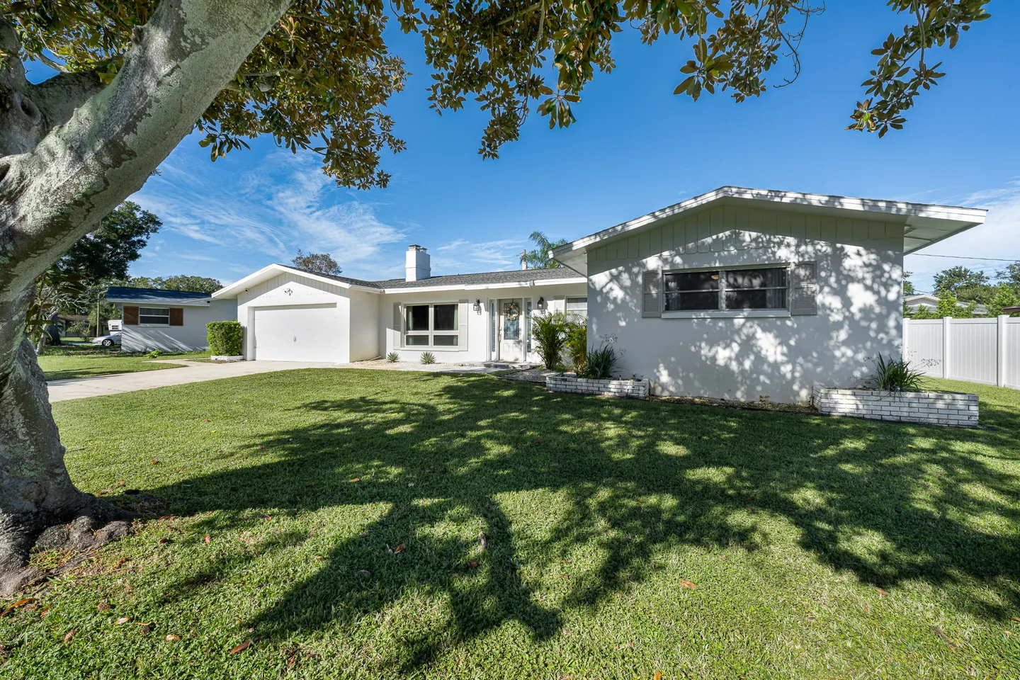 Exterior front view of a single-story white home with a green lawn, shaded by a large tree under a blue sky.