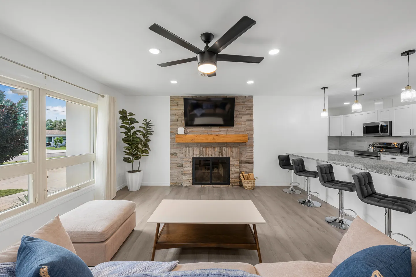 Open living area with a stone fireplace, mounted TV, light wood-look floors, and an adjacent kitchen counter with barstools.