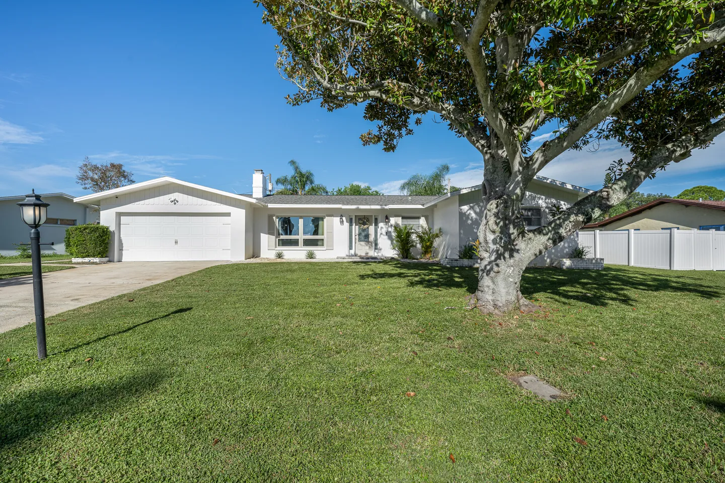 Exterior view of a single-story white house with a green lawn, large shade tree, and attached garage under a blue sky.