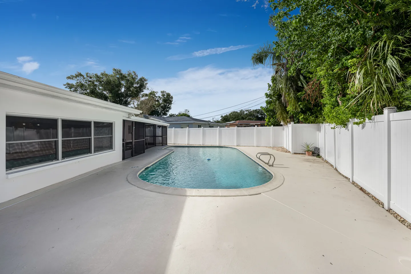 Outdoor swimming pool surrounded by a concrete patio, a screened patio entryway, and tall white privacy fencing under a blue sky.