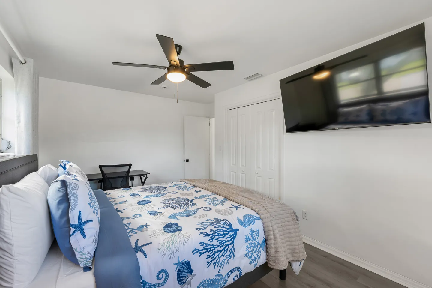 Bedroom with a bed made in blue and white coastal bedding, a ceiling fan, a wall-mounted TV, and a desk with a chair in the background.