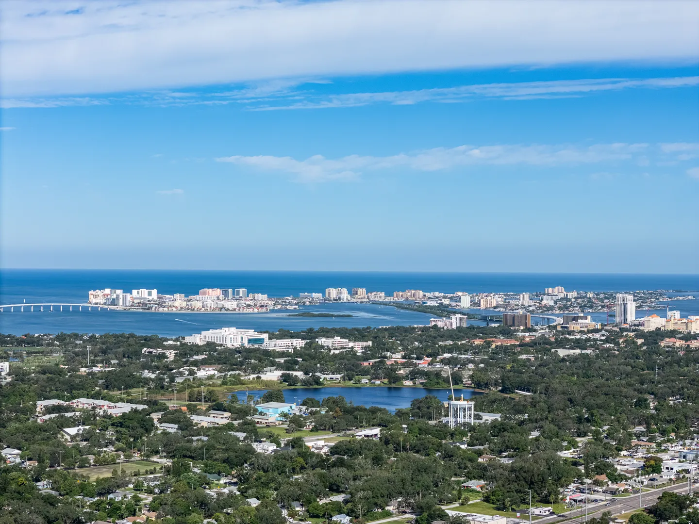 High aerial view overlooking coastal neighborhoods, inland lakes, a distant bay, bridges, and city skyline against the ocean under a blue sky.