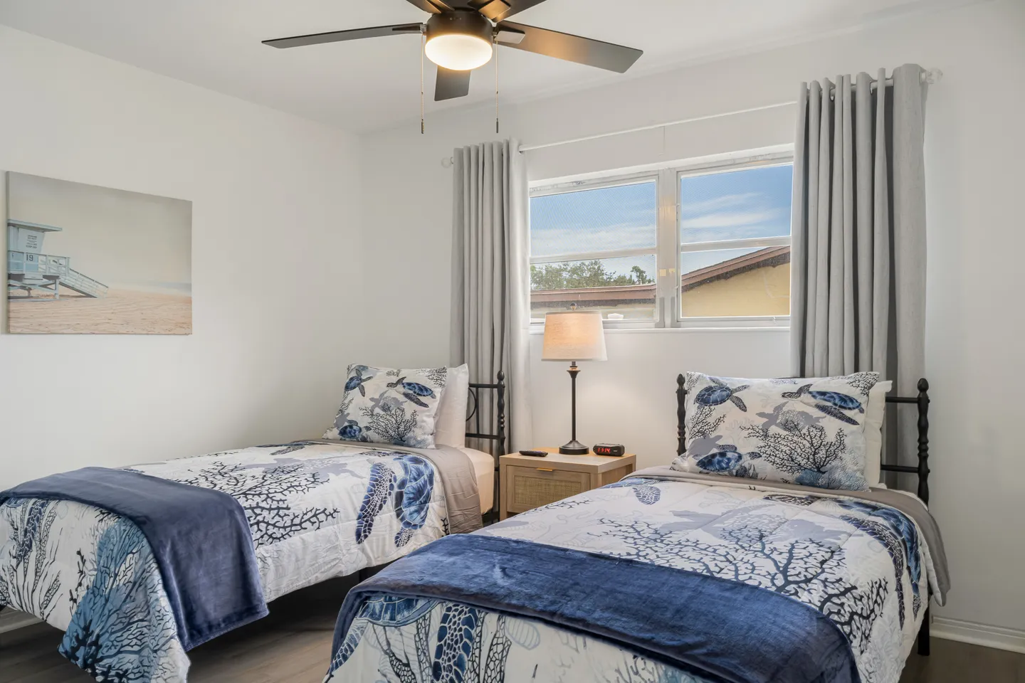 Bedroom with two twin beds dressed in coastal-themed bedding, a shared nightstand, and a window with gray curtains.