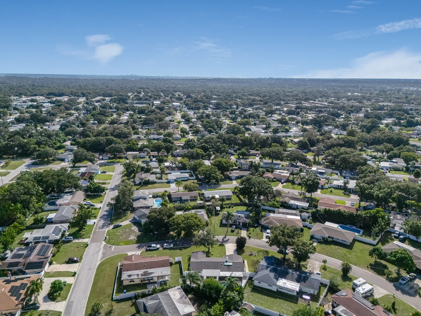 Aerial view of a residential neighborhood with single-story homes, mature green trees, and paved streets under a blue sky.
