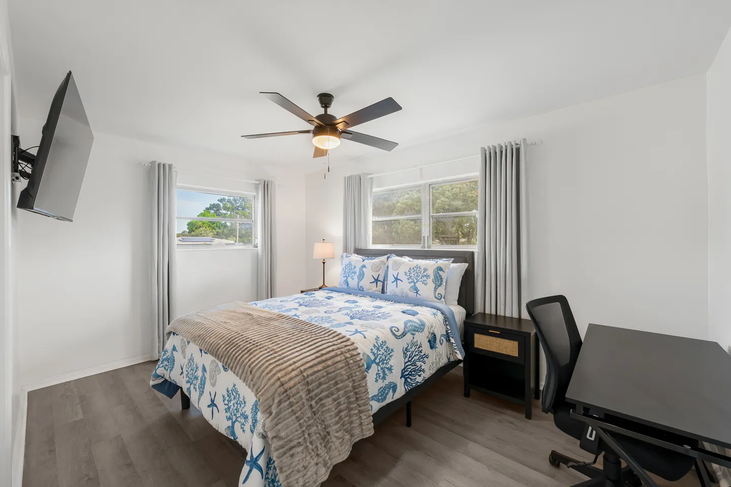 Bedroom with coastal-themed bedding, a wall-mounted TV, two windows with curtains, and a desk with an office chair.