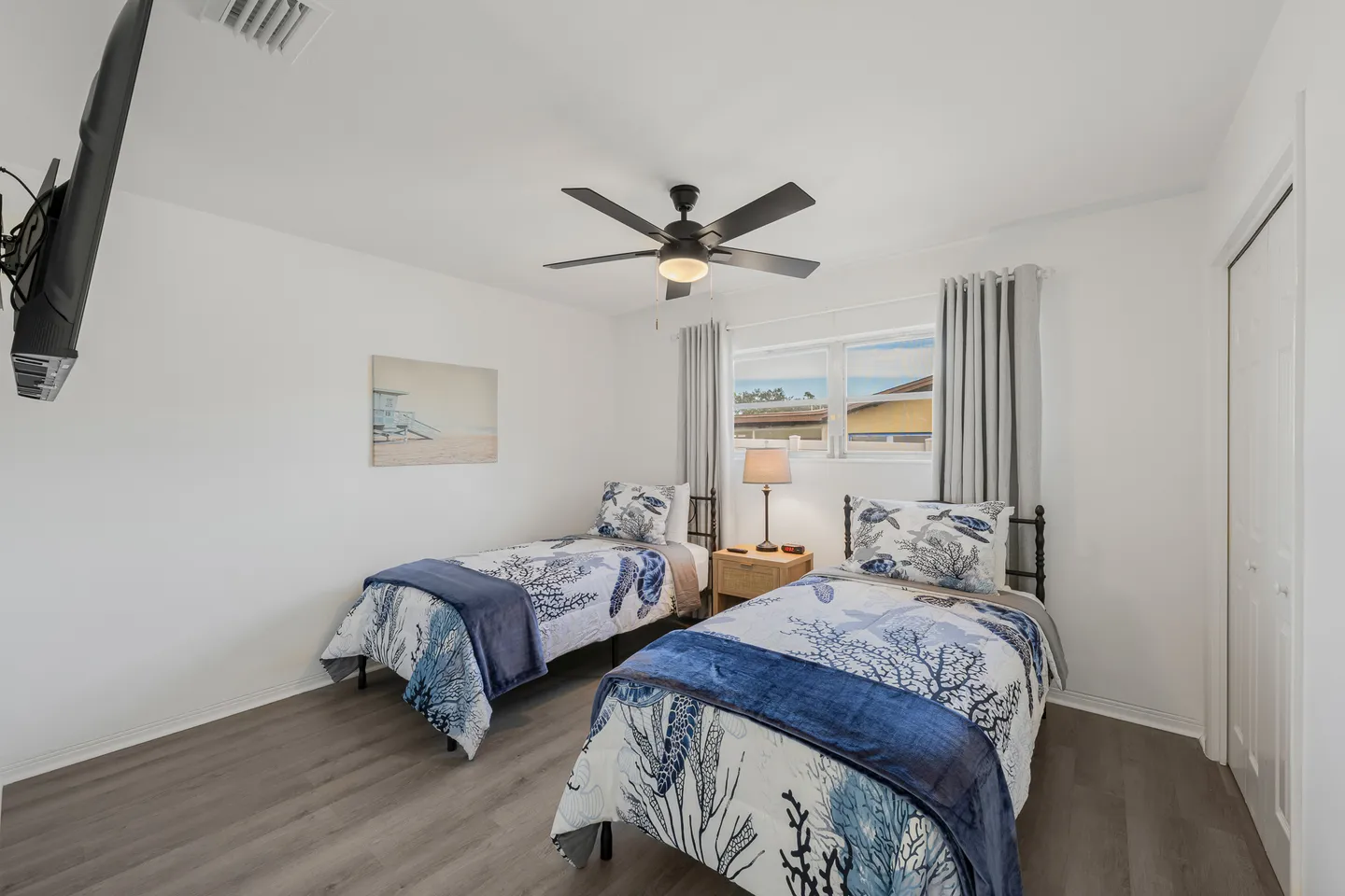 A bedroom with two twin beds featuring coastal-themed bedding, a nightstand with a lamp between them, and a ceiling fan overhead.