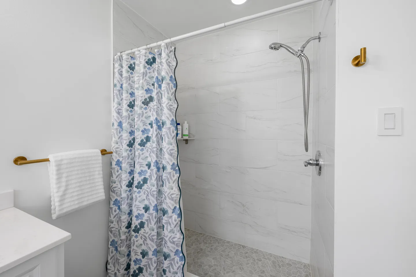 Walk-in shower with marble-look tiled walls, pebble floor, floral shower curtain, and handheld showerhead.
