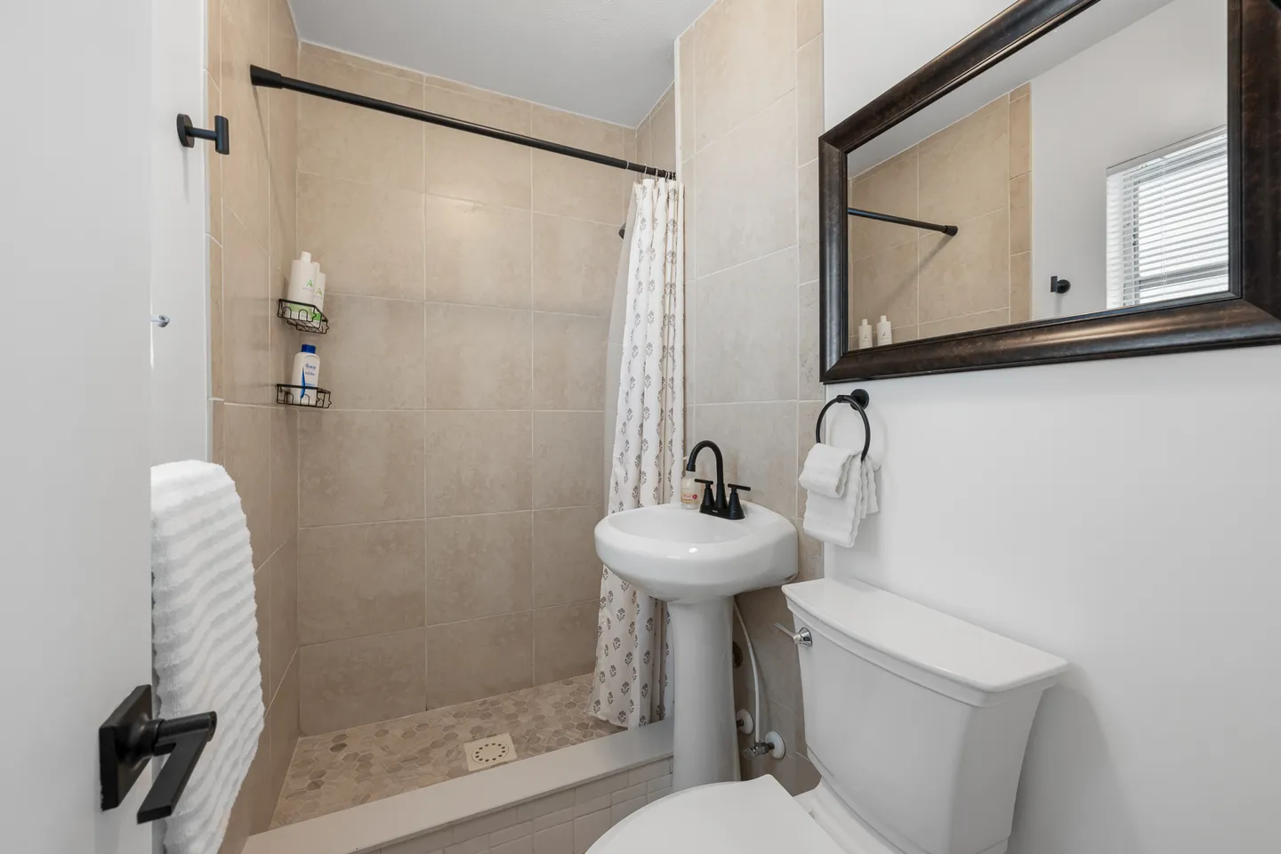 Bathroom with a pedestal sink, toilet, framed mirror, and a walk-in shower with beige tile.