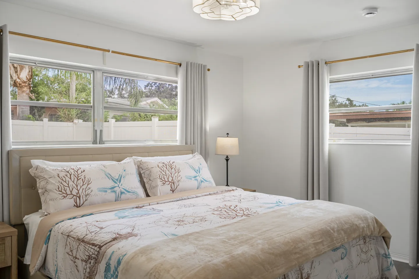 Bedroom with coastal-themed bedding, an upholstered headboard, a nightstand with a lamp, and windows looking outside.