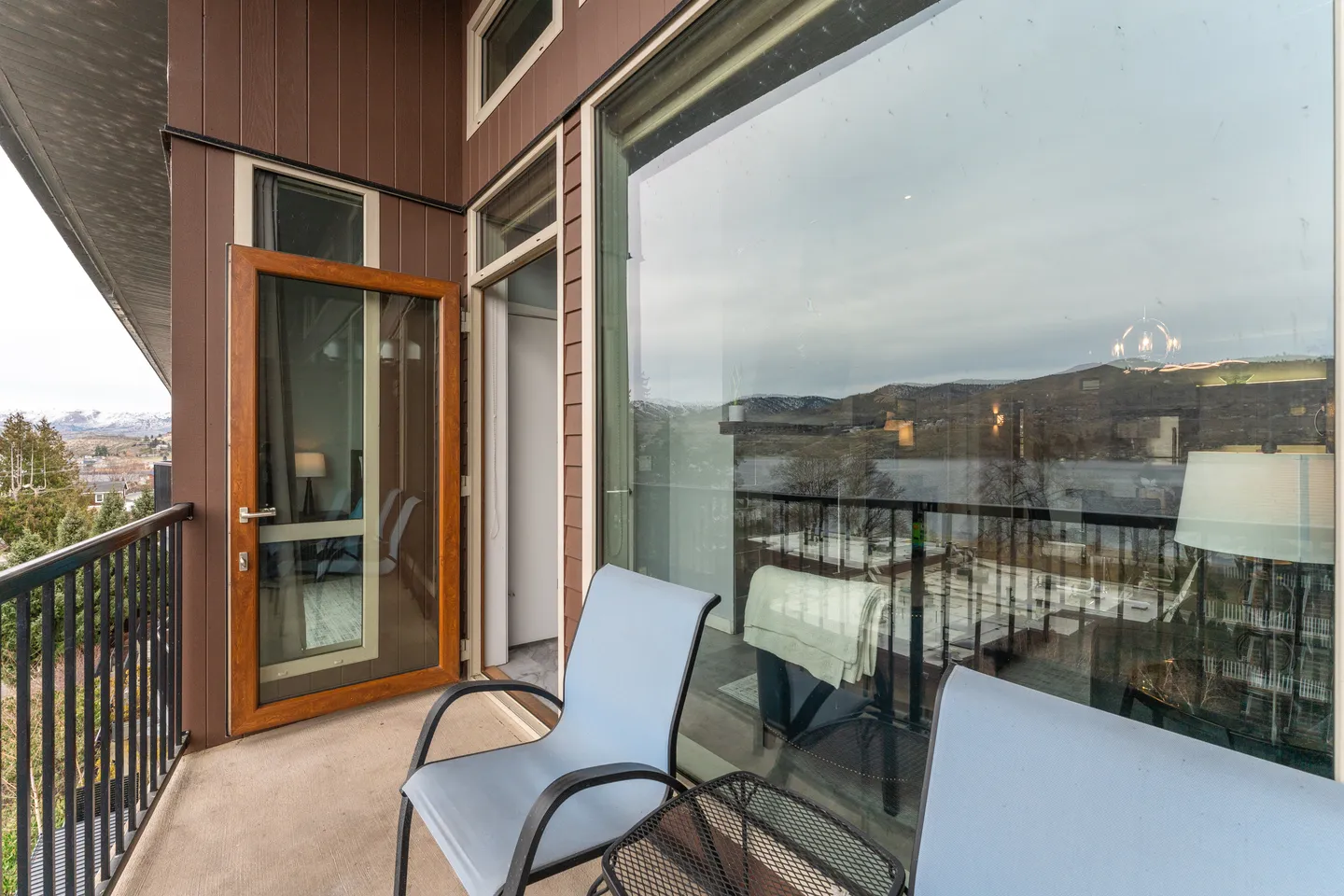 Balcony with two light blue chairs, a small table, dark railings, and a large window reflecting the mountain landscape.