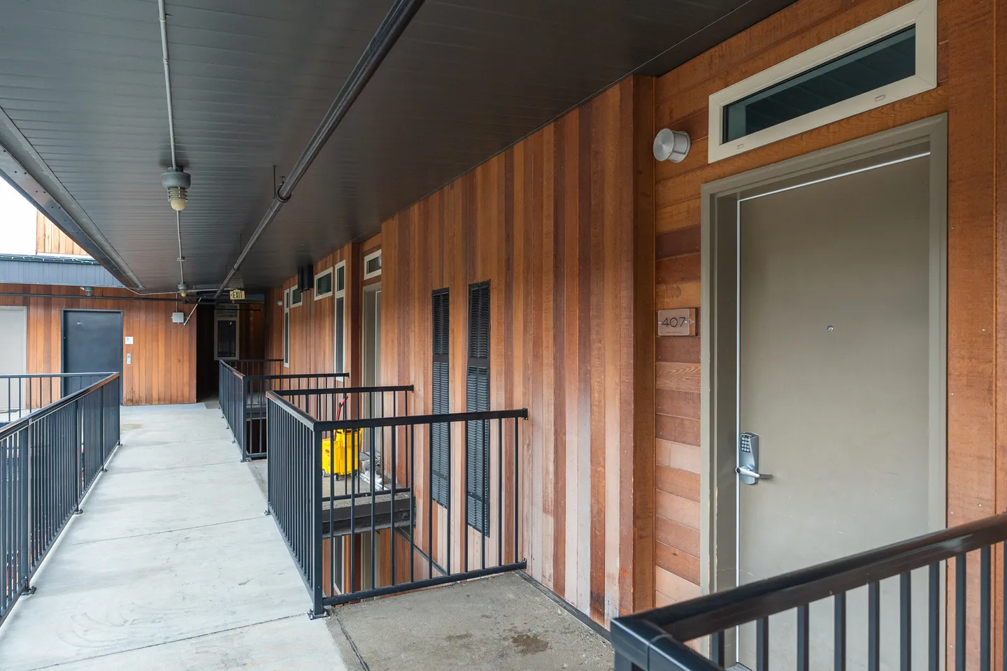 Exterior open-air hallway of a residential building with vertical wood siding, metal railings, and entrance doors.