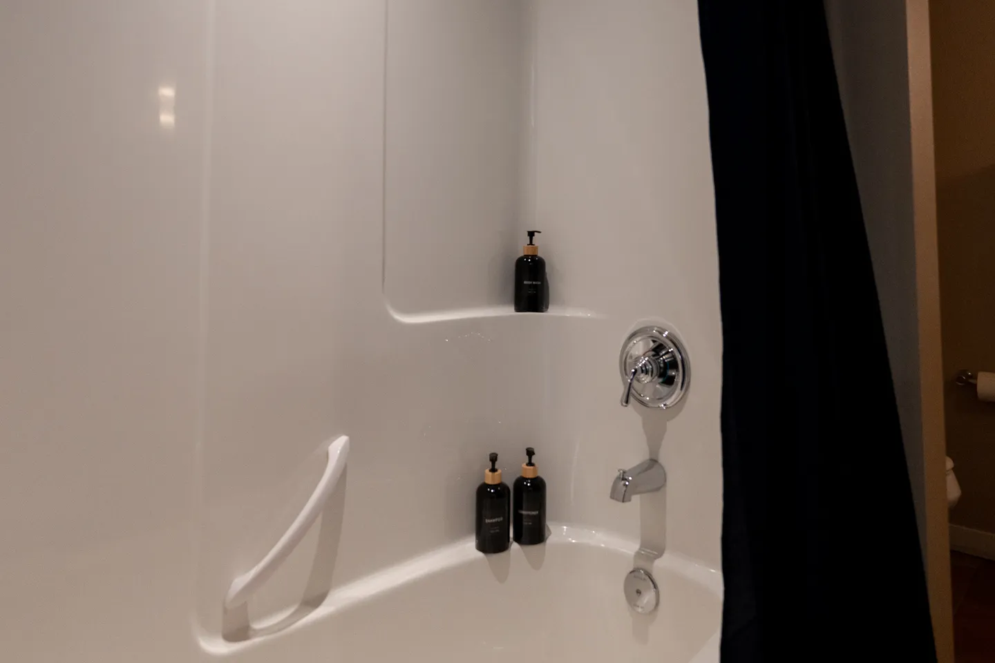 Close-up view of a white bathtub shower combination with chrome fixtures, toiletries on the built-in shelves, and a dark shower curtain.