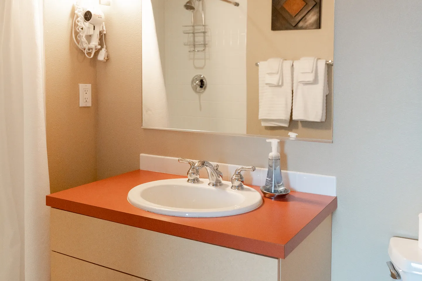 Bathroom vanity with an oval sink, red-orange countertop, mirror, wall-mounted hairdryer, and reflected shower.