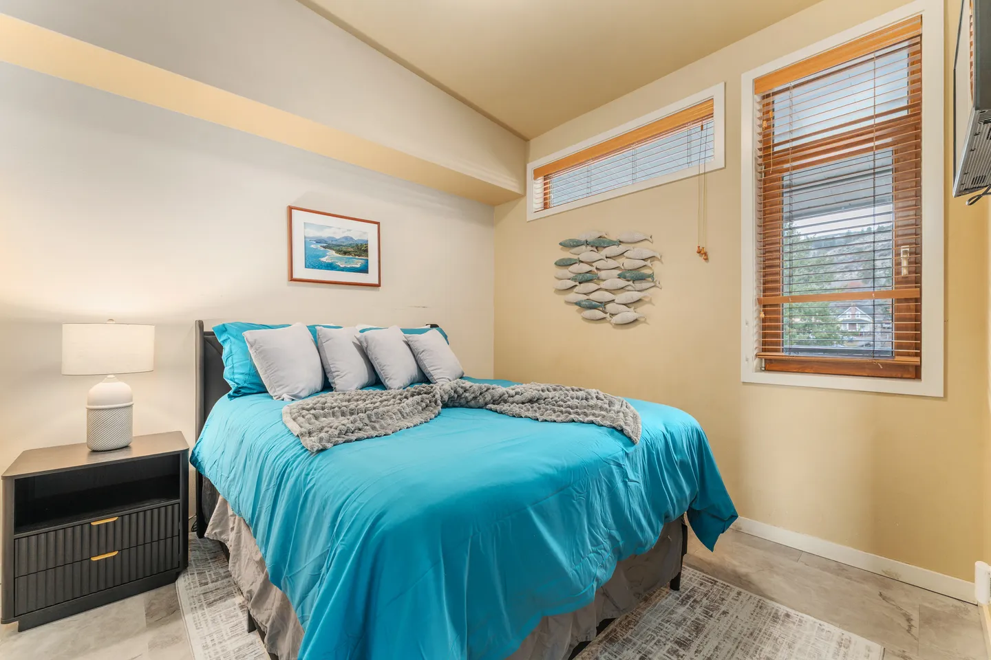 Bedroom with a bed made in bright blue bedding, a black nightstand with a lamp, and windows with wooden blinds.