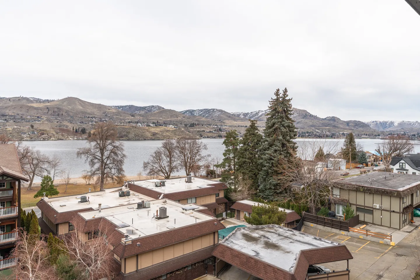 Elevated view overlooking residential buildings toward a large lake and mountains under an overcast sky.