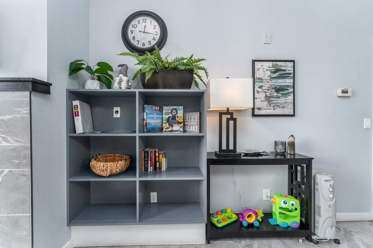 A gray six-cube shelving unit holding books and a woven basket, next to a black side table with a lamp and toys.