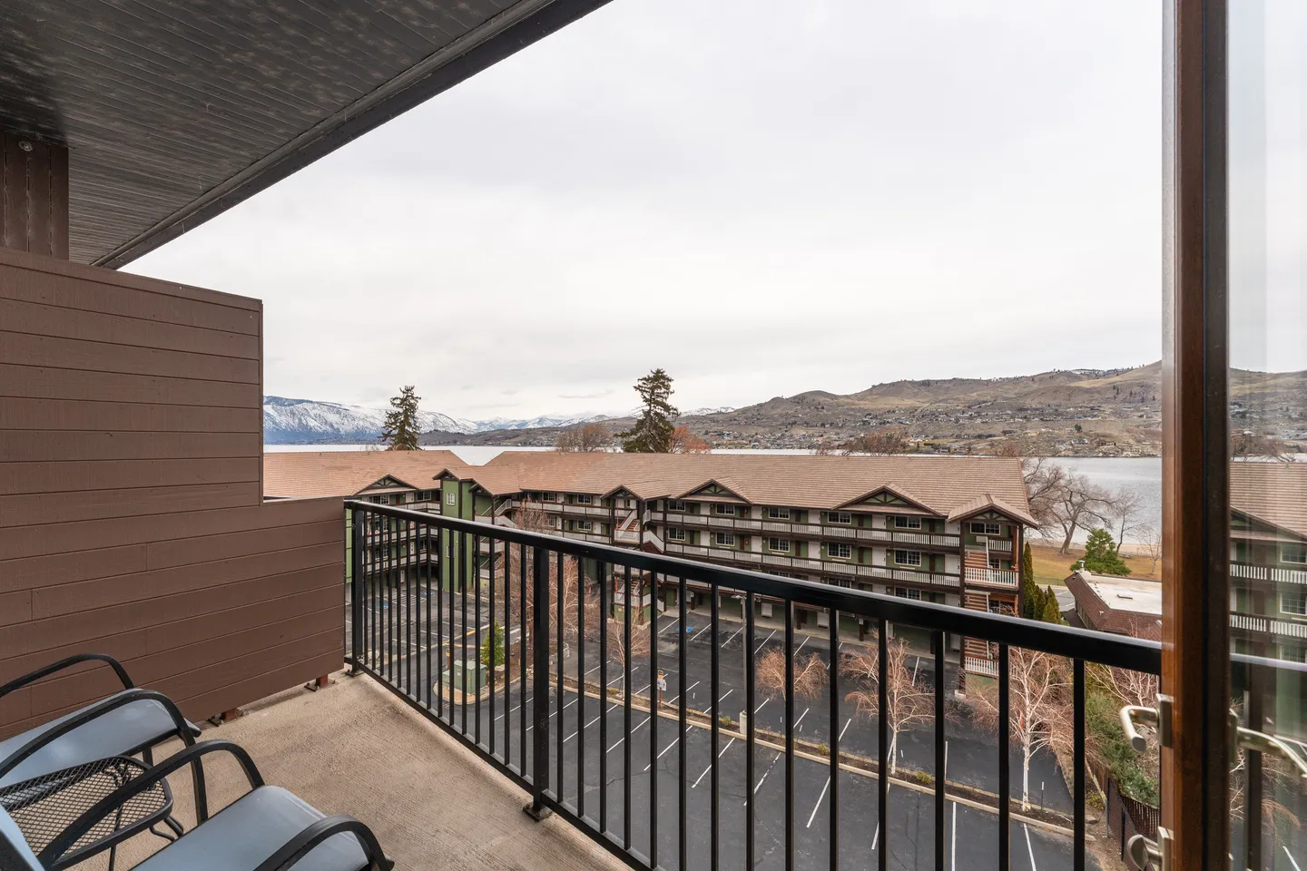 Balcony view overlooking a parking lot, adjacent resort buildings, a lake, and snow-dusted mountains under an overcast sky.