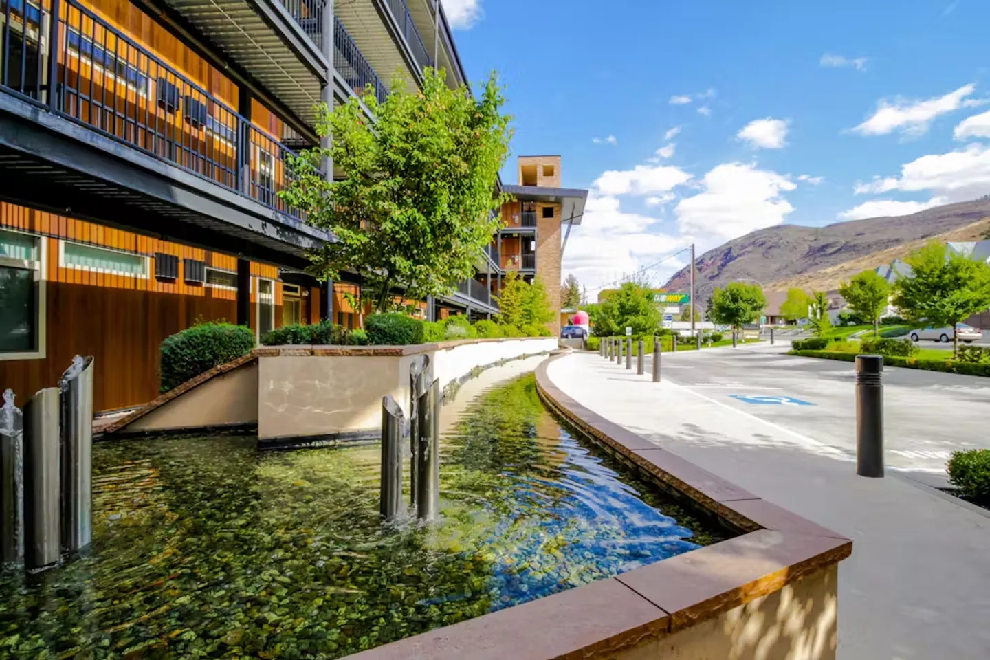 Exterior view of a multi-story building featuring a stepped water fountain with fountains spraying into a clear stone-lined pool along a paved walkway.