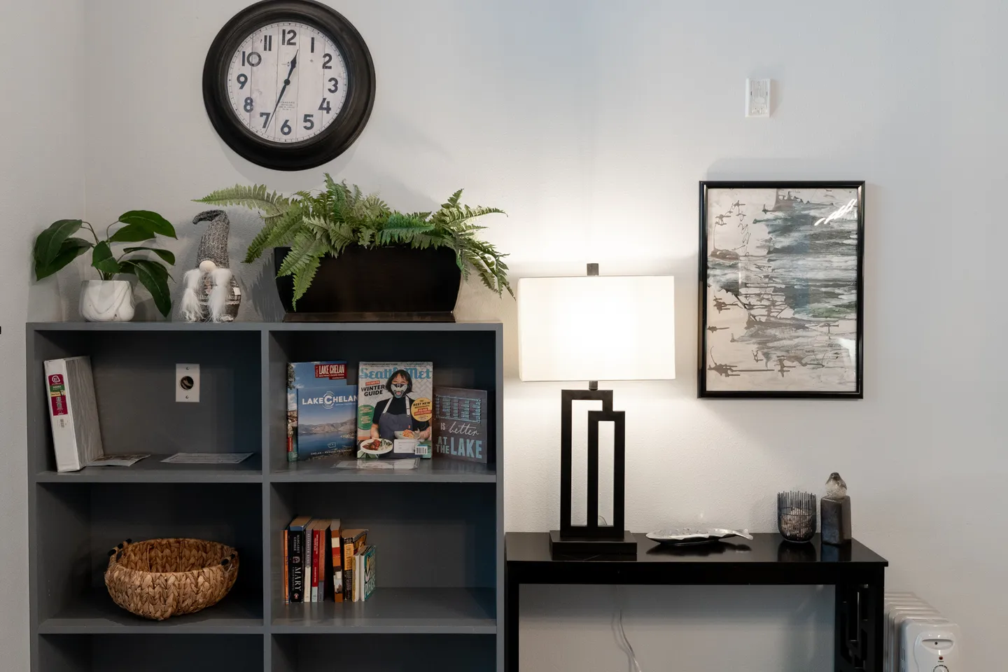 A gray cubby bookcase with books, plants, and decor next to a black side table with a lit lamp.