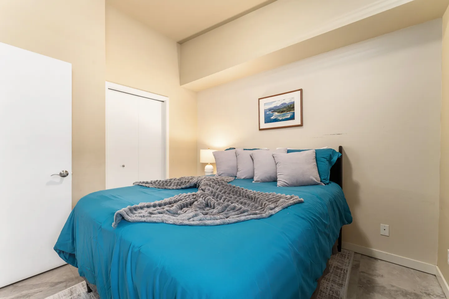 Bedroom with a bed made in blue linens and accent pillows, a bedside lamp, and a white closet door.