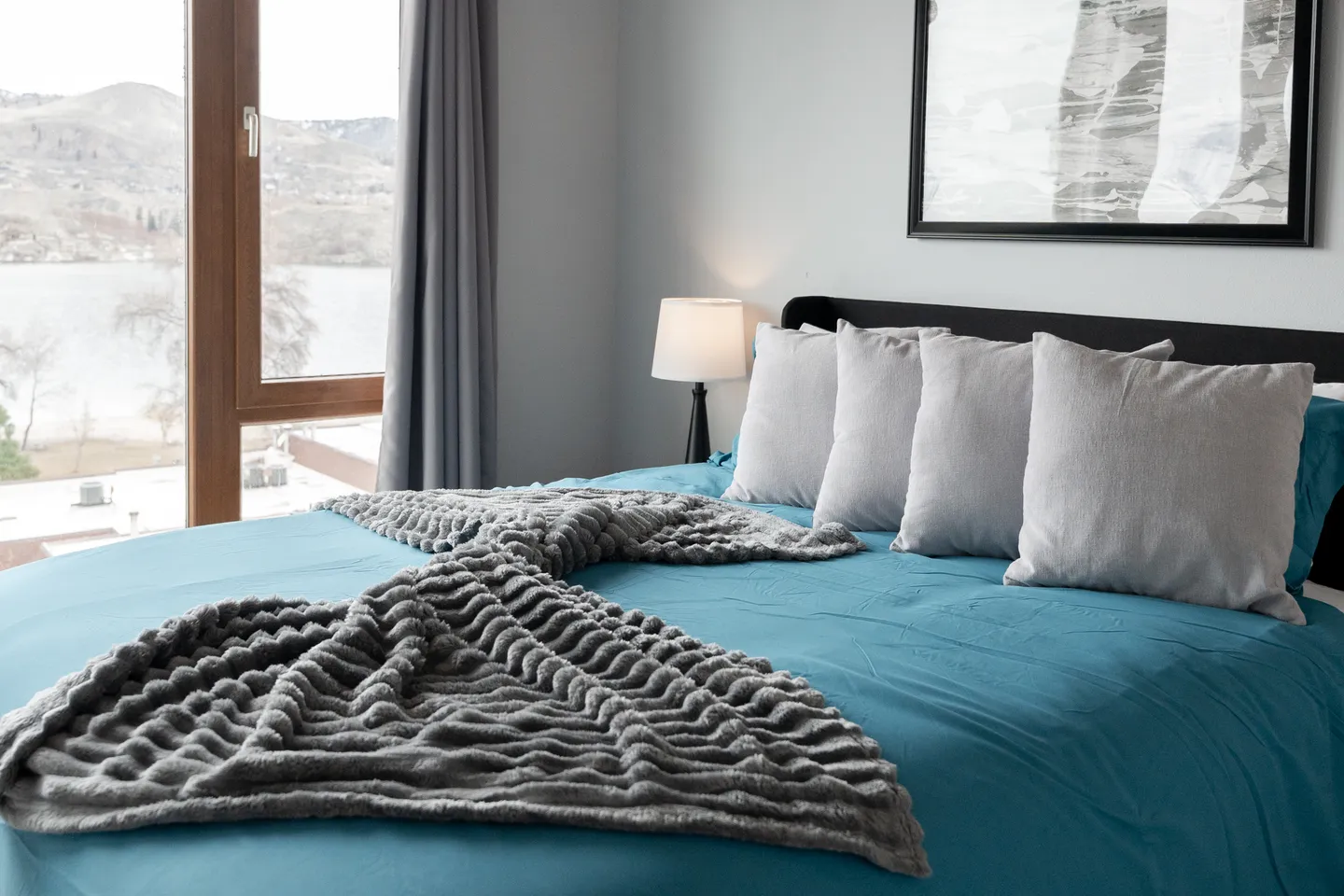Bedroom with a bed made in blue and grey linens next to a window overlooking water and hills.