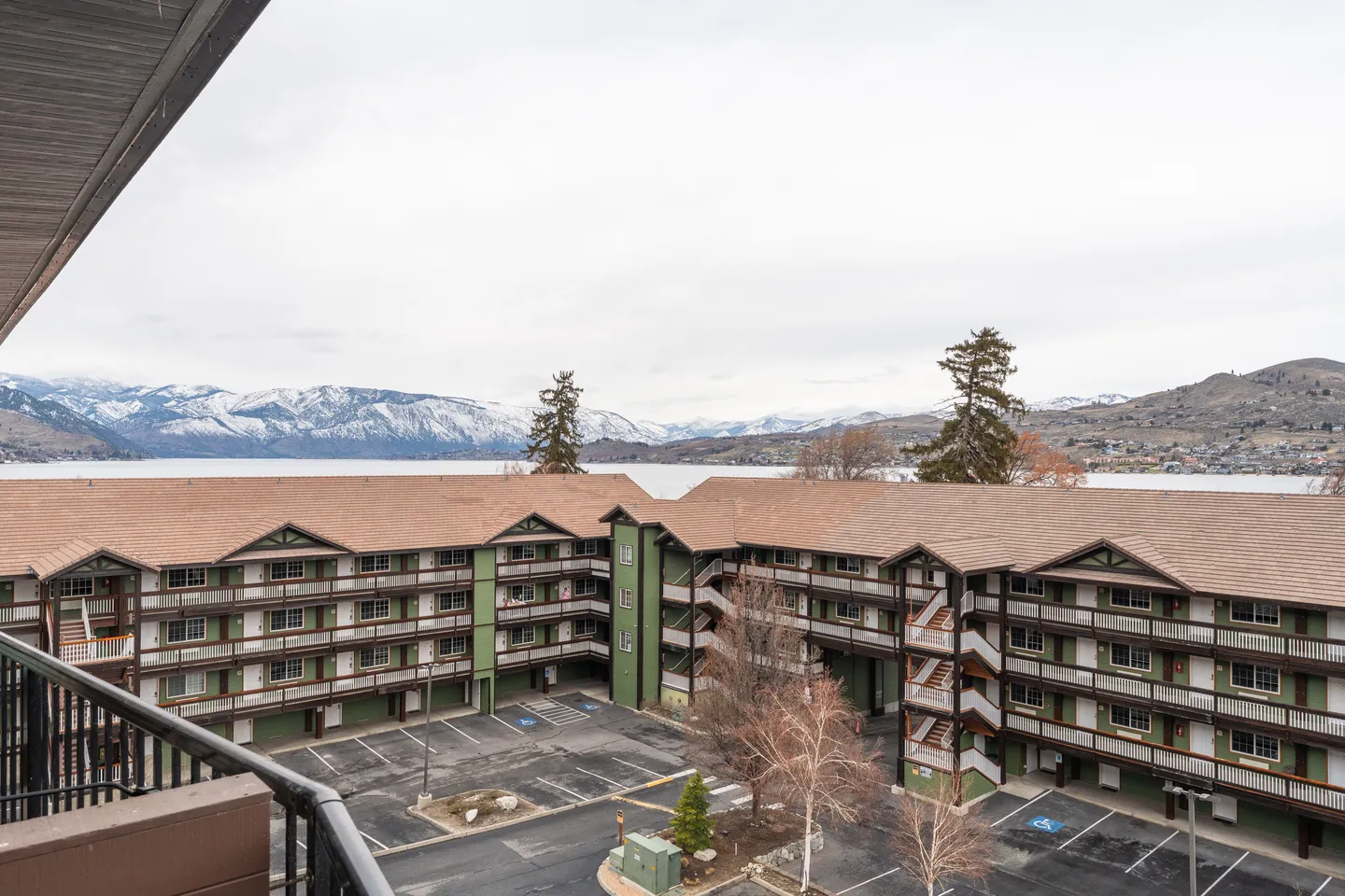 Balcony view overlooking a multi-story condominium complex, parking lot, lake, and snow-dusted mountains under an overcast sky.