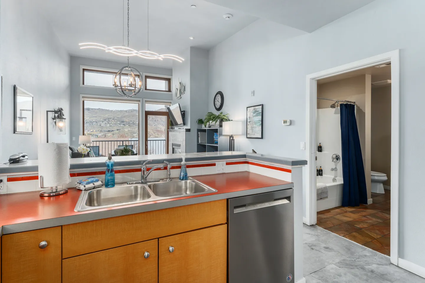 Kitchen counter and sink looking through to an open living room with large windows and an adjacent bathroom.