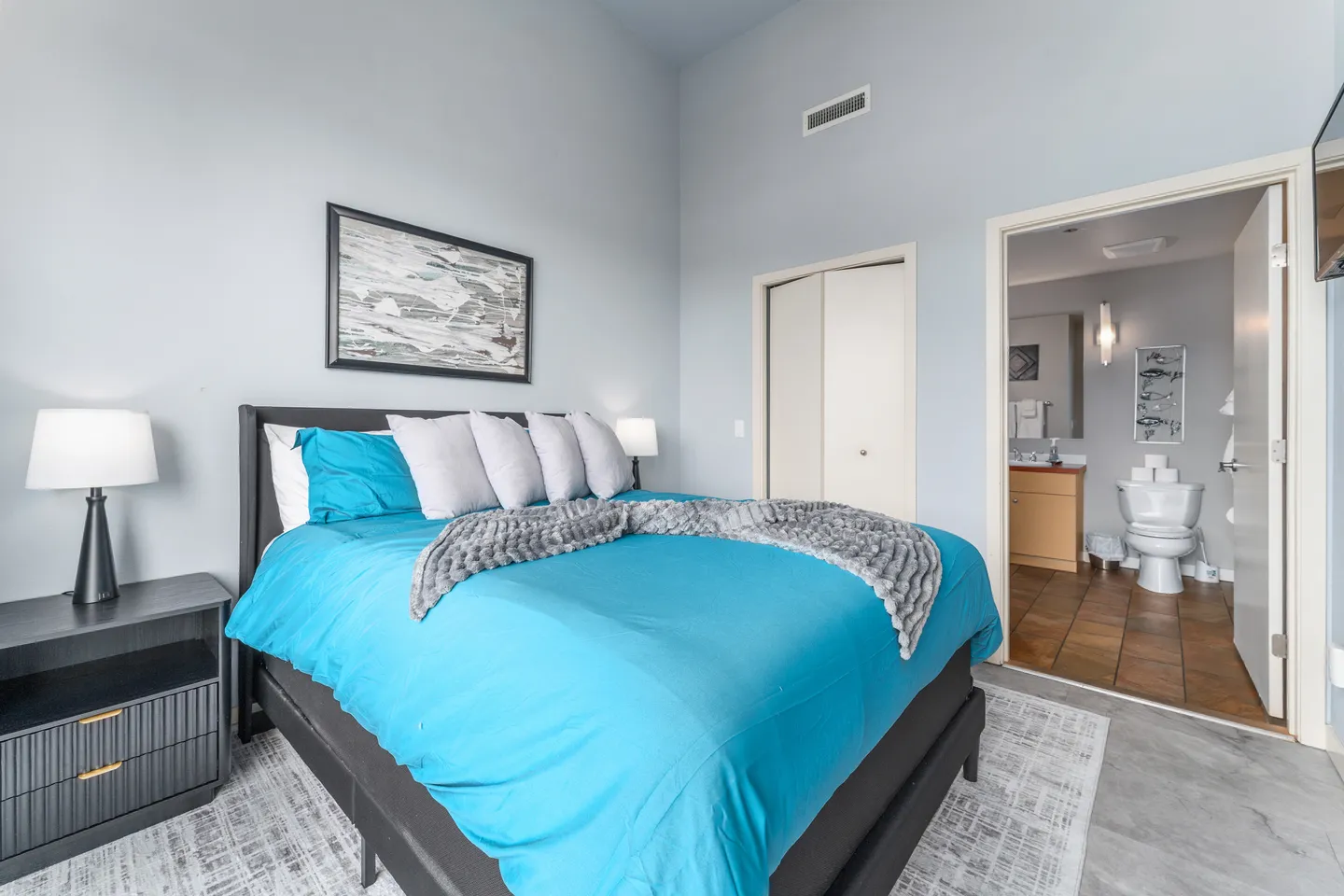 Bedroom with a turquoise-dressed bed, dark nightstand, closet doors, and an open doorway leading into an en-suite bathroom.