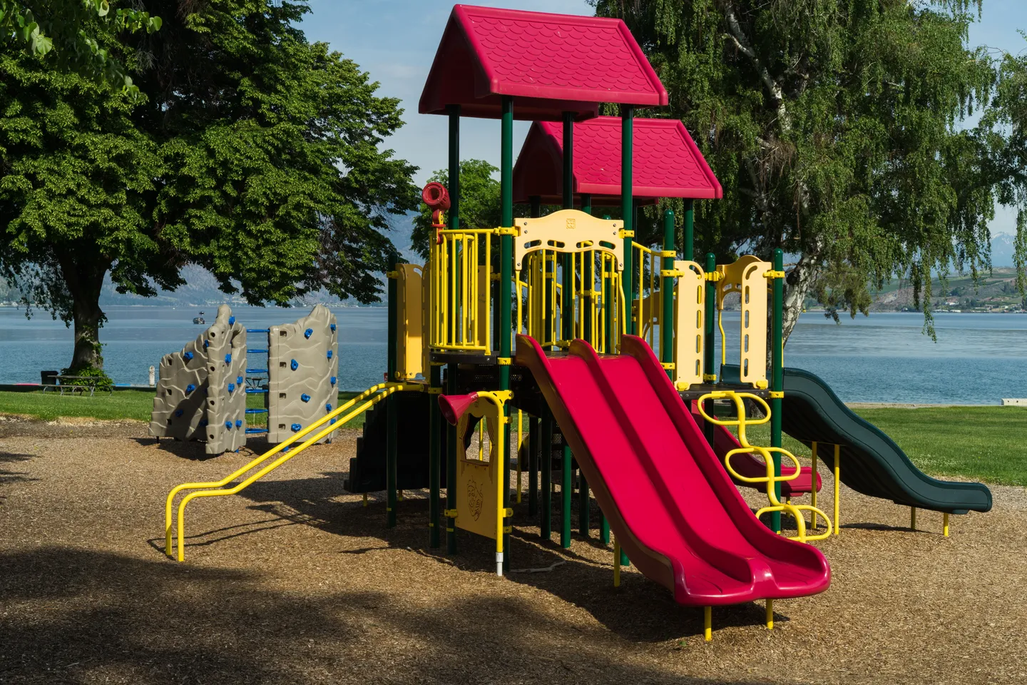 Outdoor children's playground structure with red slides next to a lake surrounded by green trees.