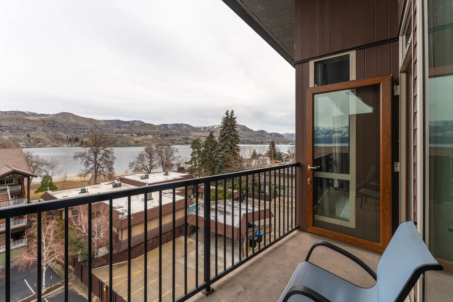 Balcony with black metal railings and a blue chair overlooking a lake, buildings, and distant hills under an overcast sky.