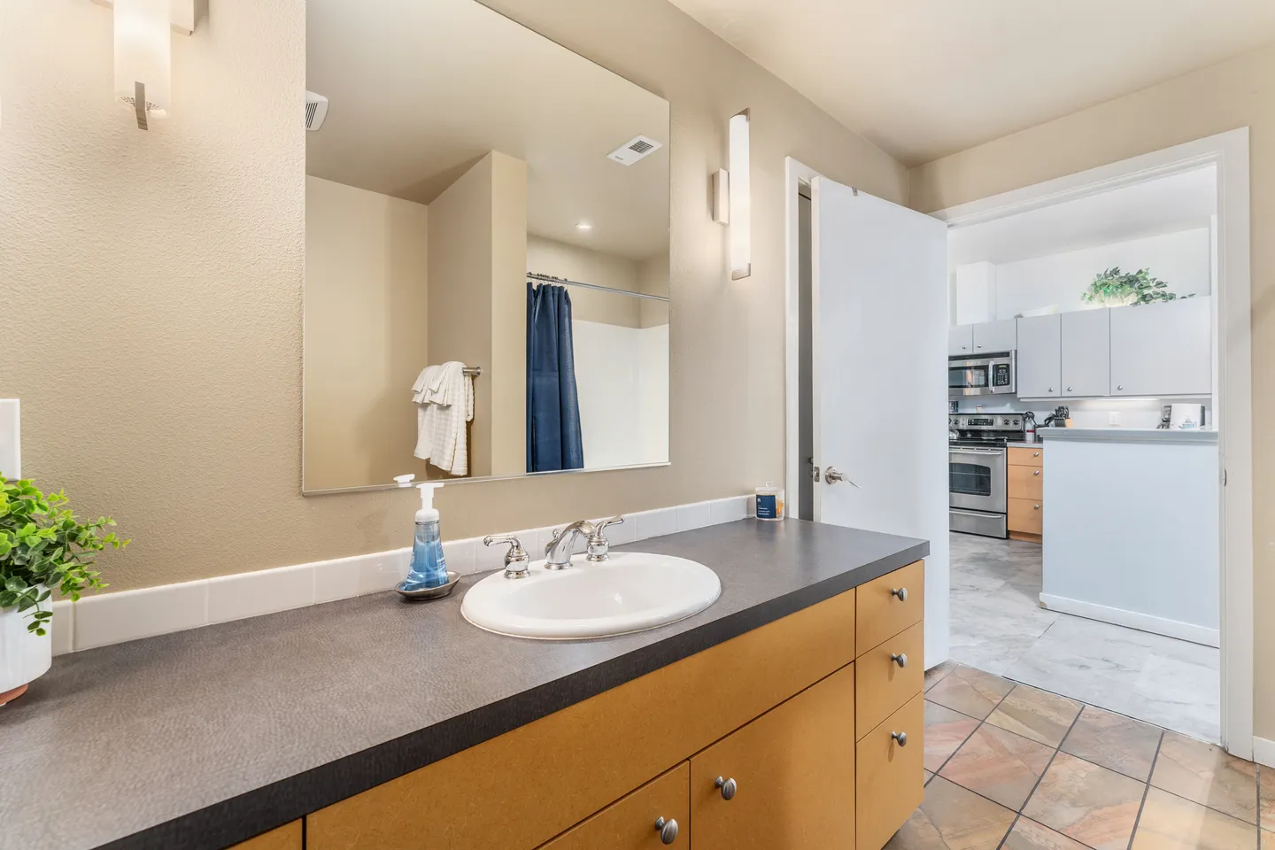 Bathroom vanity with dark countertop and oval sink, reflecting a shower curtain in the large mirror, with an open doorway leading toward the kitchen.