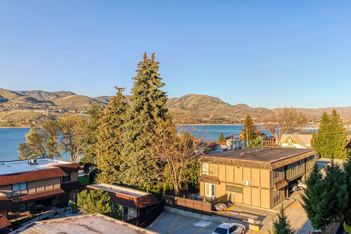 Elevated view overlooking residential buildings, evergreen trees, and a lake with rolling hills in the background.