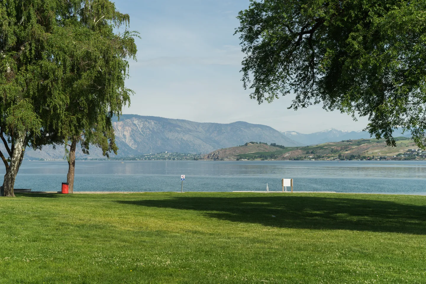 Grassy waterfront park overlooking a wide lake with mountains in the background under clear skies.