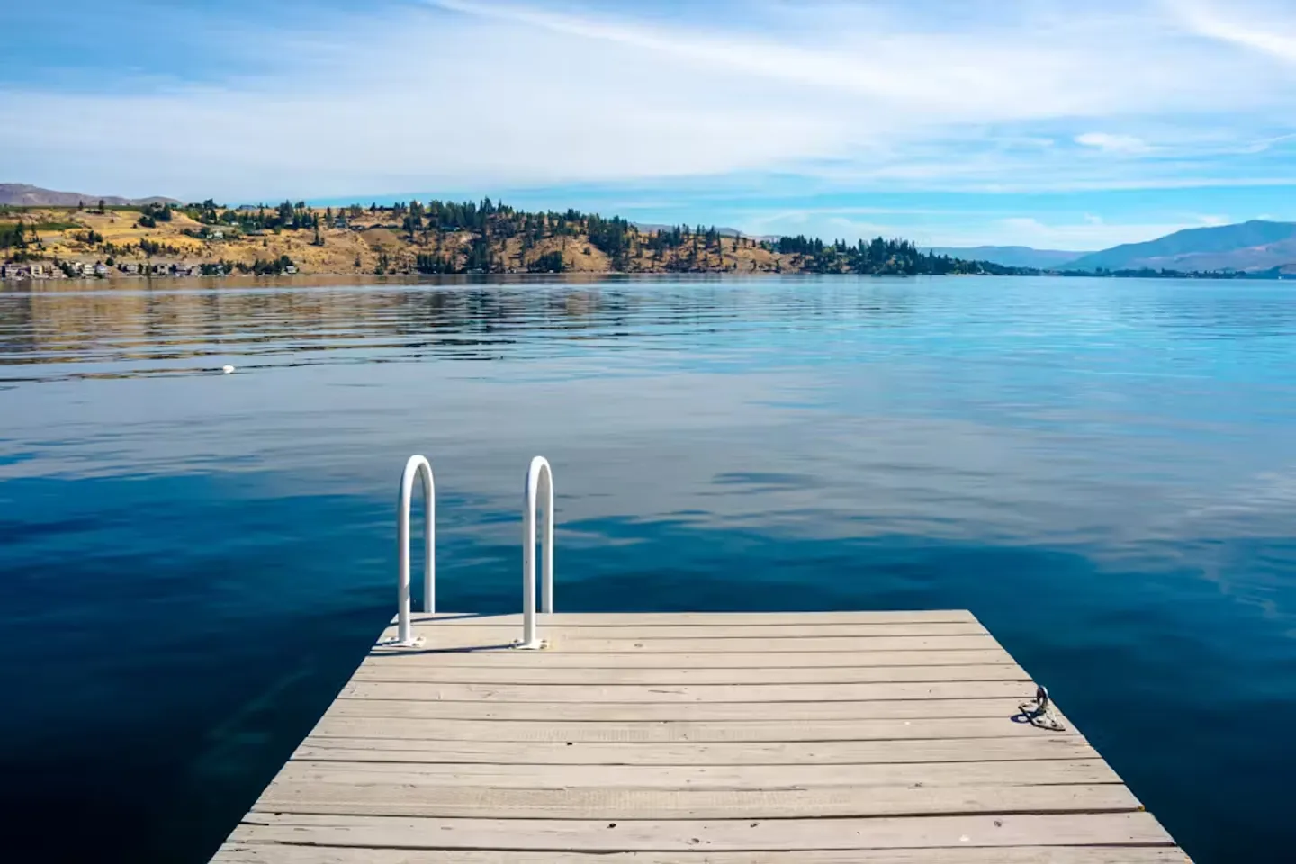 A wooden dock with a white swim ladder extending into a calm blue lake, with hills across the water under a partly cloudy sky.