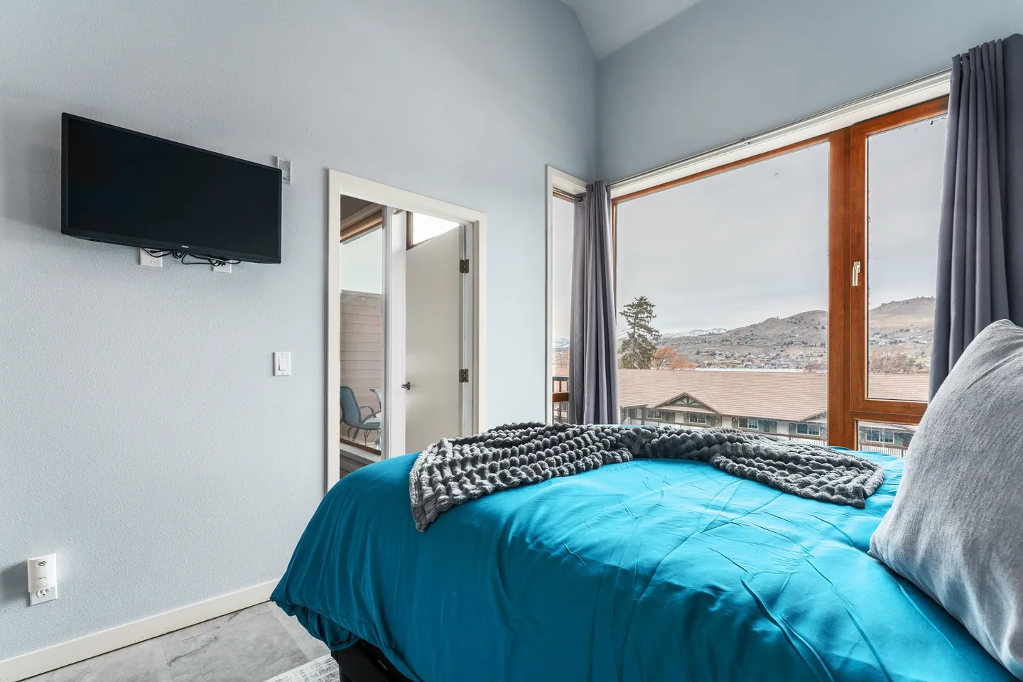 Bedroom with a bed made with a bright blue comforter next to a large picture window overlooking hills.