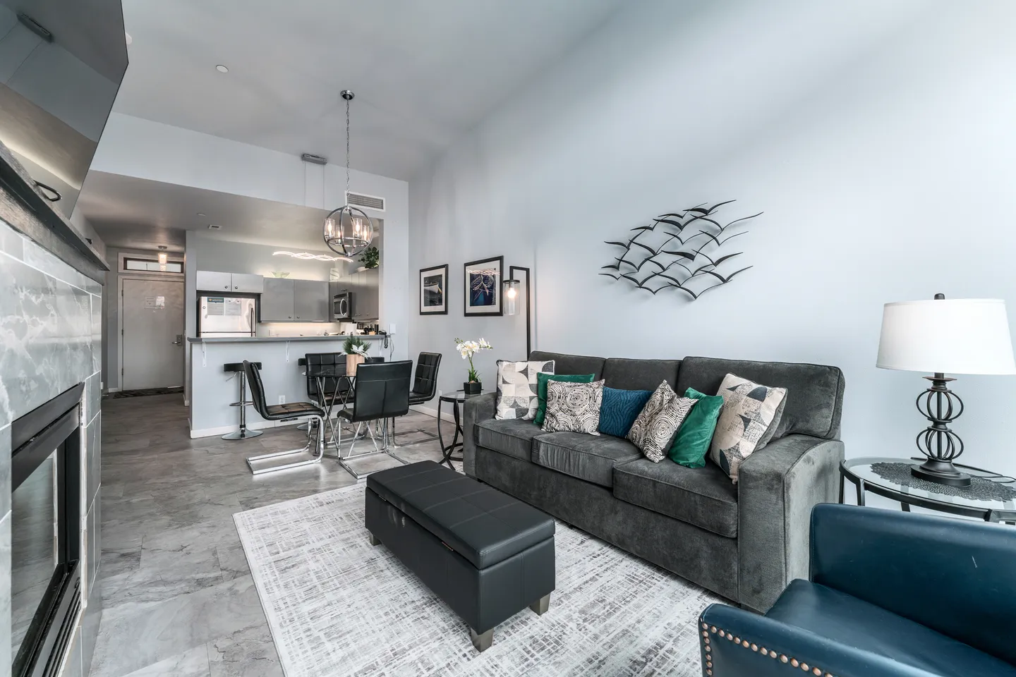 Open-concept living room featuring a grey sofa, ottoman, and fireplace, leading to a dining area and kitchen.