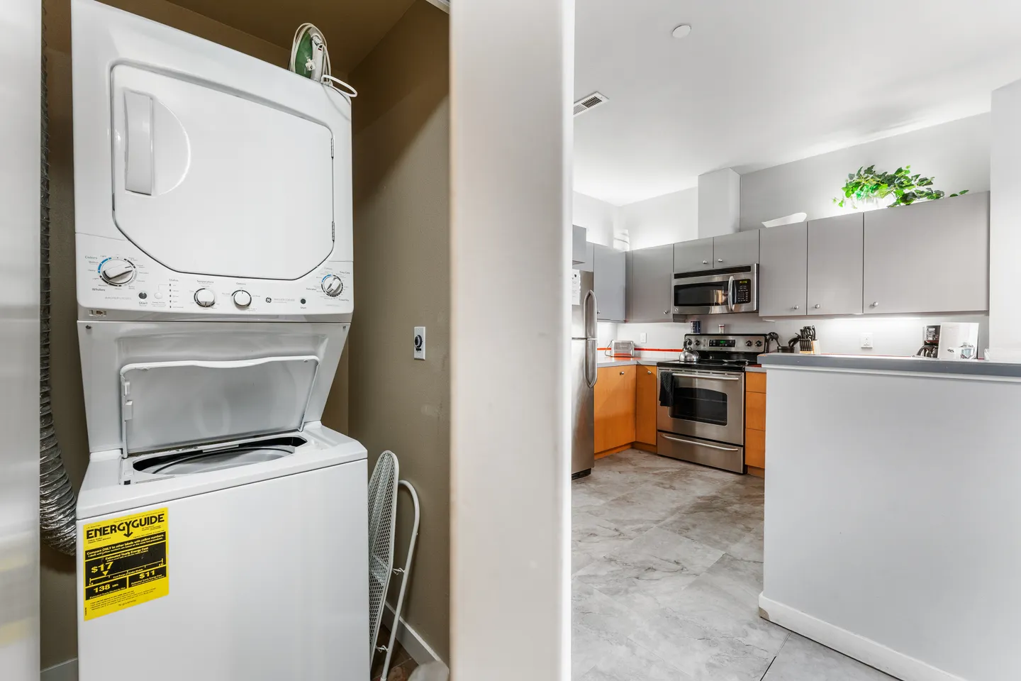 A stacked white washer and dryer unit in a closet opening, with a view into an adjacent kitchen with stainless steel appliances.