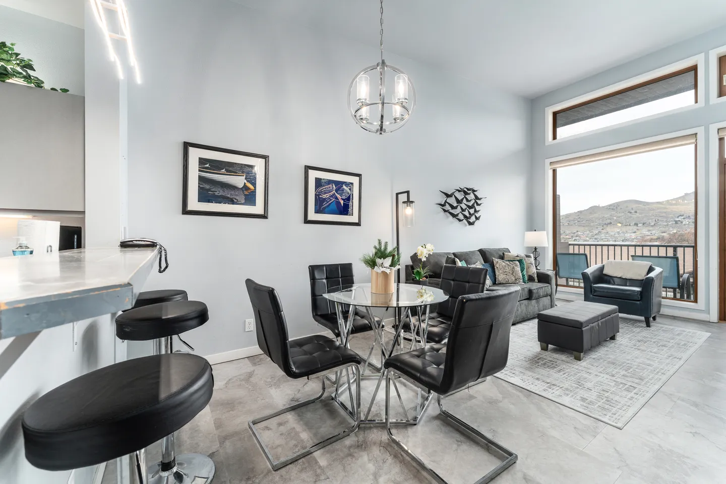Open-concept living and dining area with high ceilings, a glass-top table with four chairs, counter seating, and large windows.