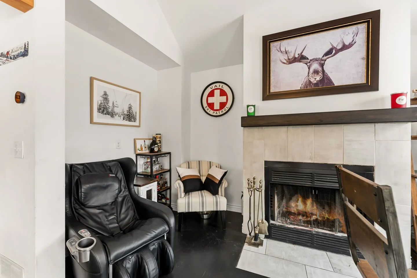 Living room corner featuring a tiled fireplace with an active fire, framed moose artwork above the mantel, a black massage chair, and an accent armchair.