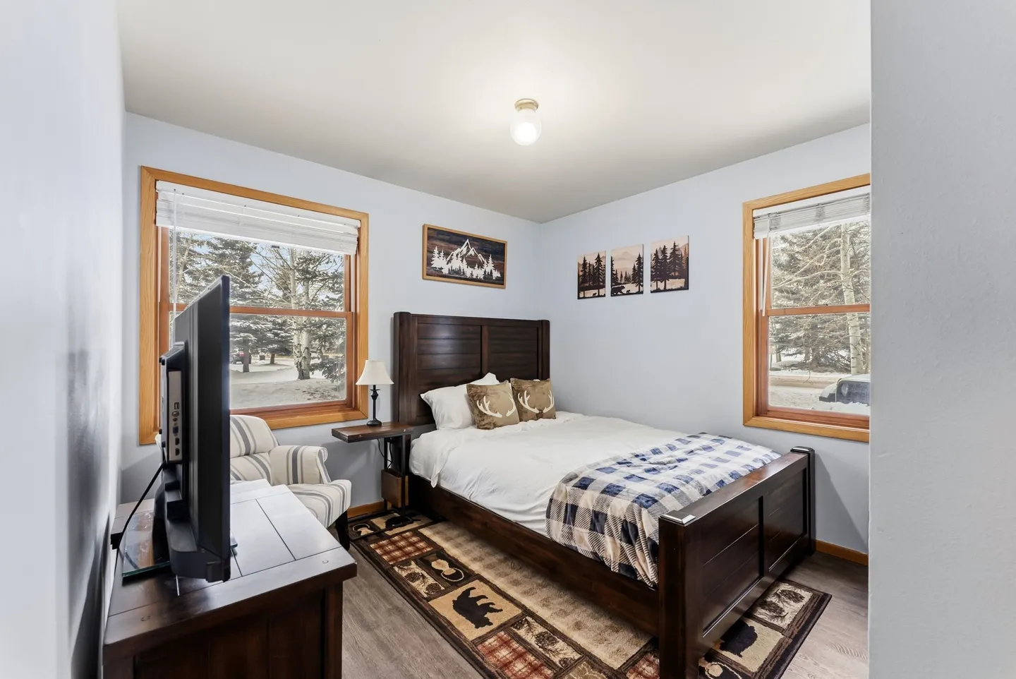 Bedroom with a dark wood bed frame, patterned area rug, armchair, and windows showing trees outside.