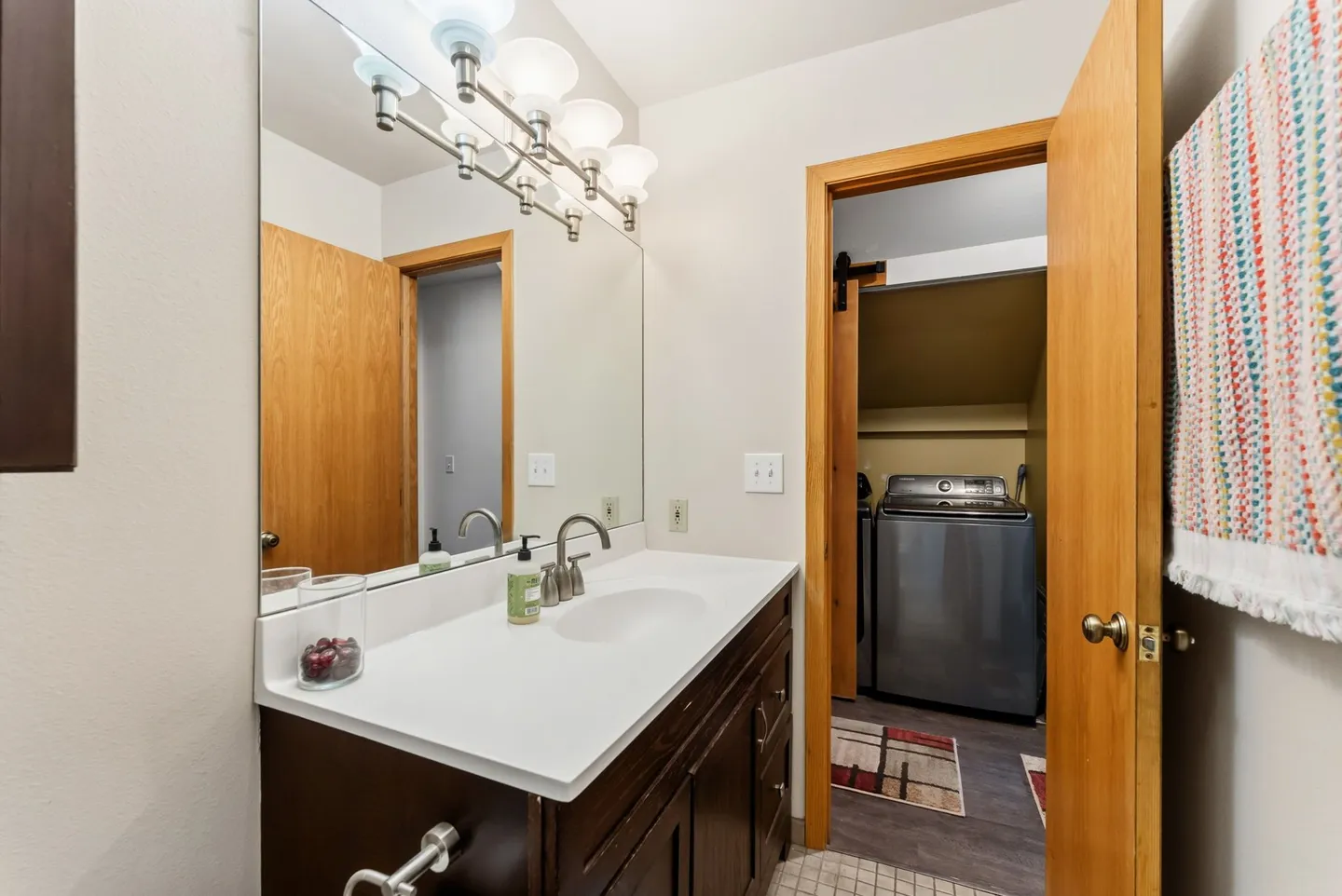 Bathroom vanity with a white countertop, dark wood cabinets, large mirror with vanity lights, and an open doorway leading to a laundry area with a washing machine.