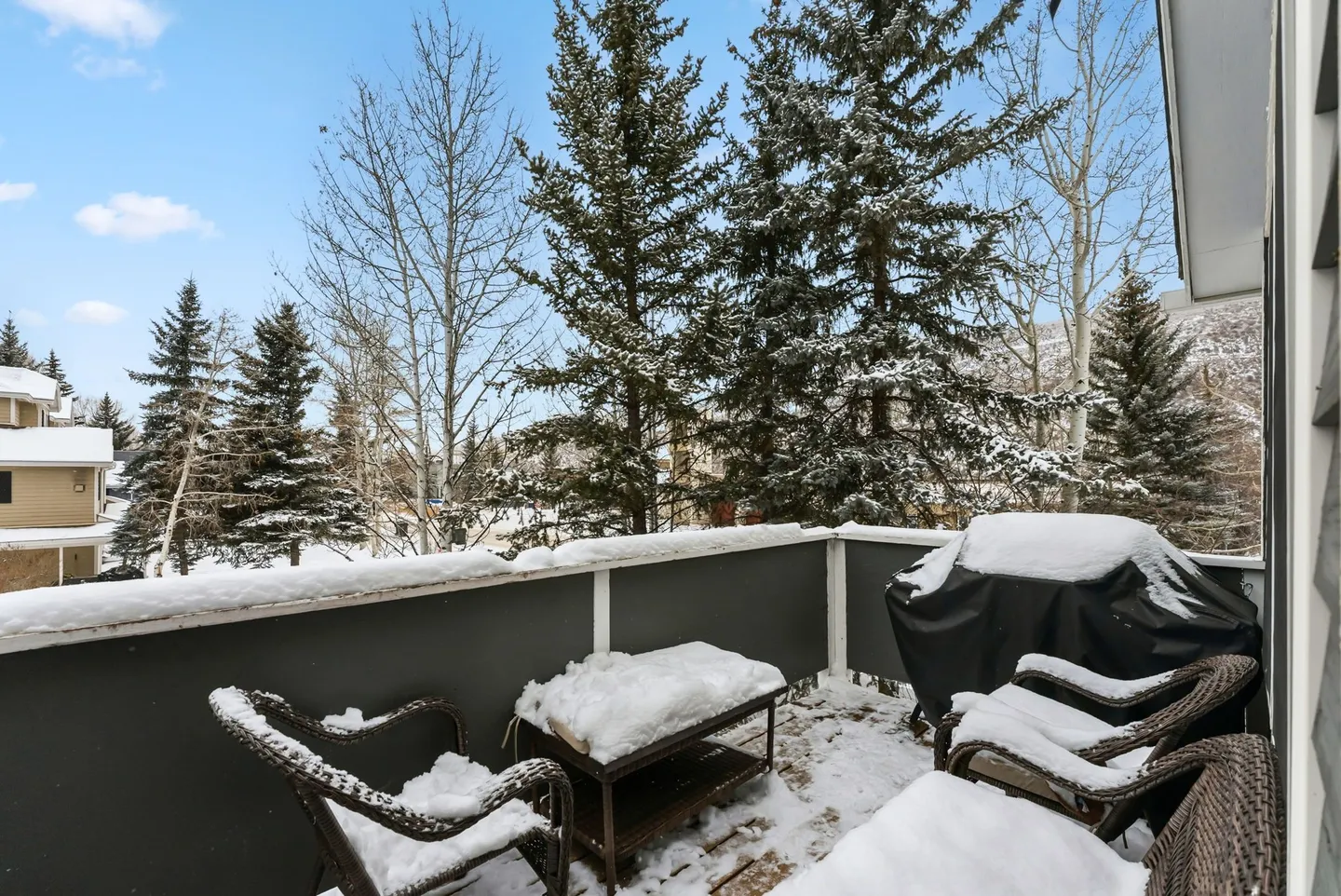 A snow-covered outdoor balcony with wicker patio chairs, a coffee table, and a covered grill overlooking pine trees.