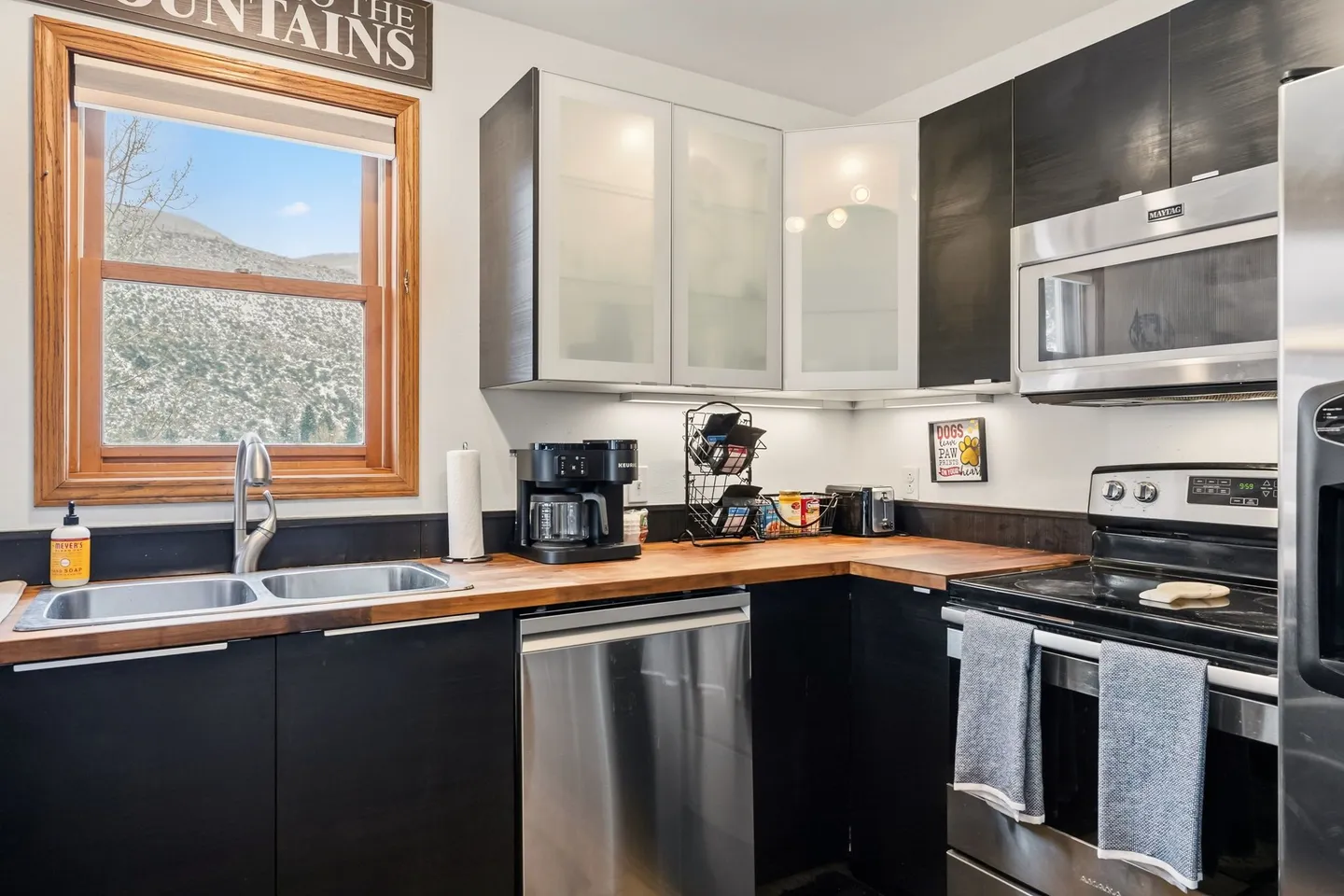 Kitchen with dark modern cabinets, butcher block countertops, stainless steel appliances, and a window overlooking mountains.