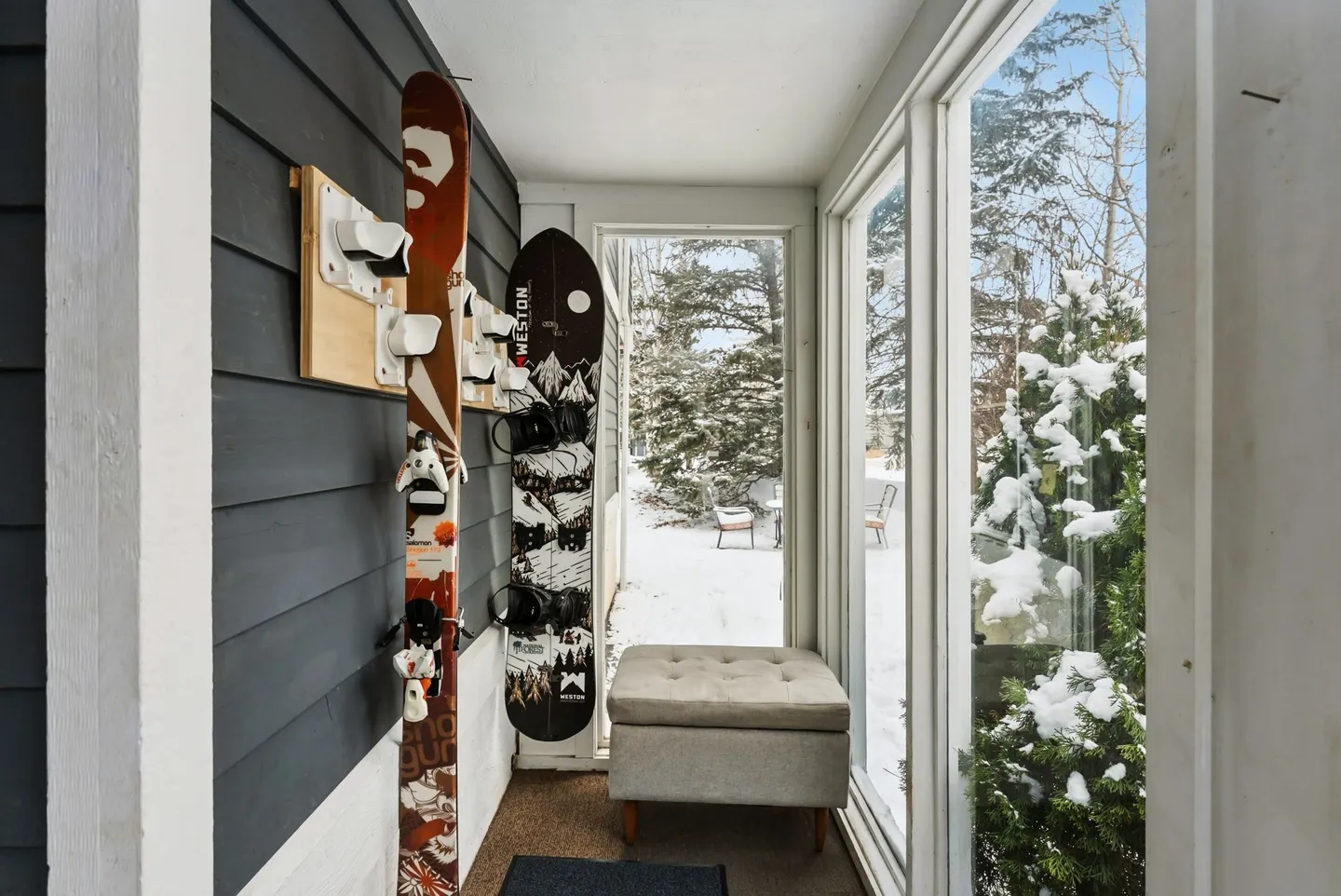 Sunroom entryway with ski and snowboard wall rack, cushioned ottoman bench, and windows looking out onto snow-covered trees.