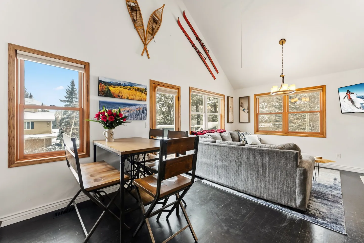 Open-concept living and dining area with vaulted ceilings, snowshoes and skis mounted on the wall, and multiple wood-framed windows.