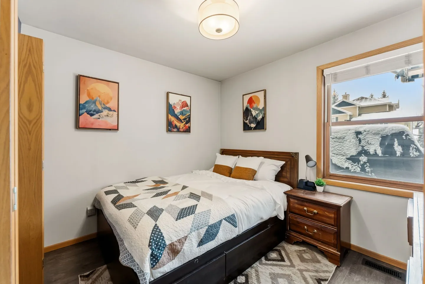 Bedroom featuring a made bed with a patterned quilt, a wooden nightstand, three framed mountain prints, and a window showing snowy outdoors.