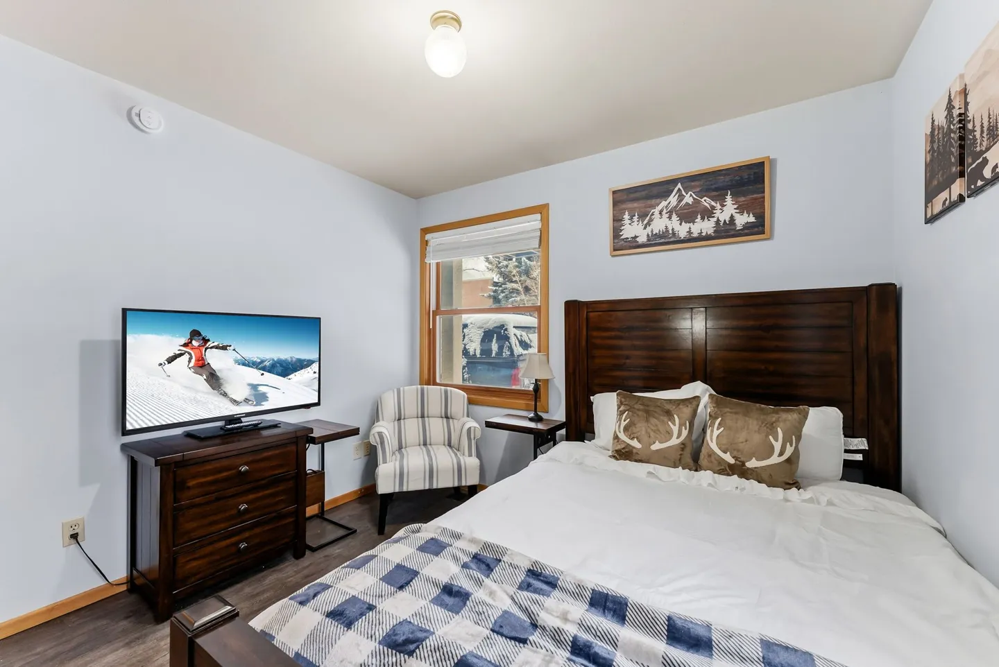 Bedroom with a dark wood bed frame, a flat-screen television on a dresser, an armchair by a window, and mountain-themed wall art.