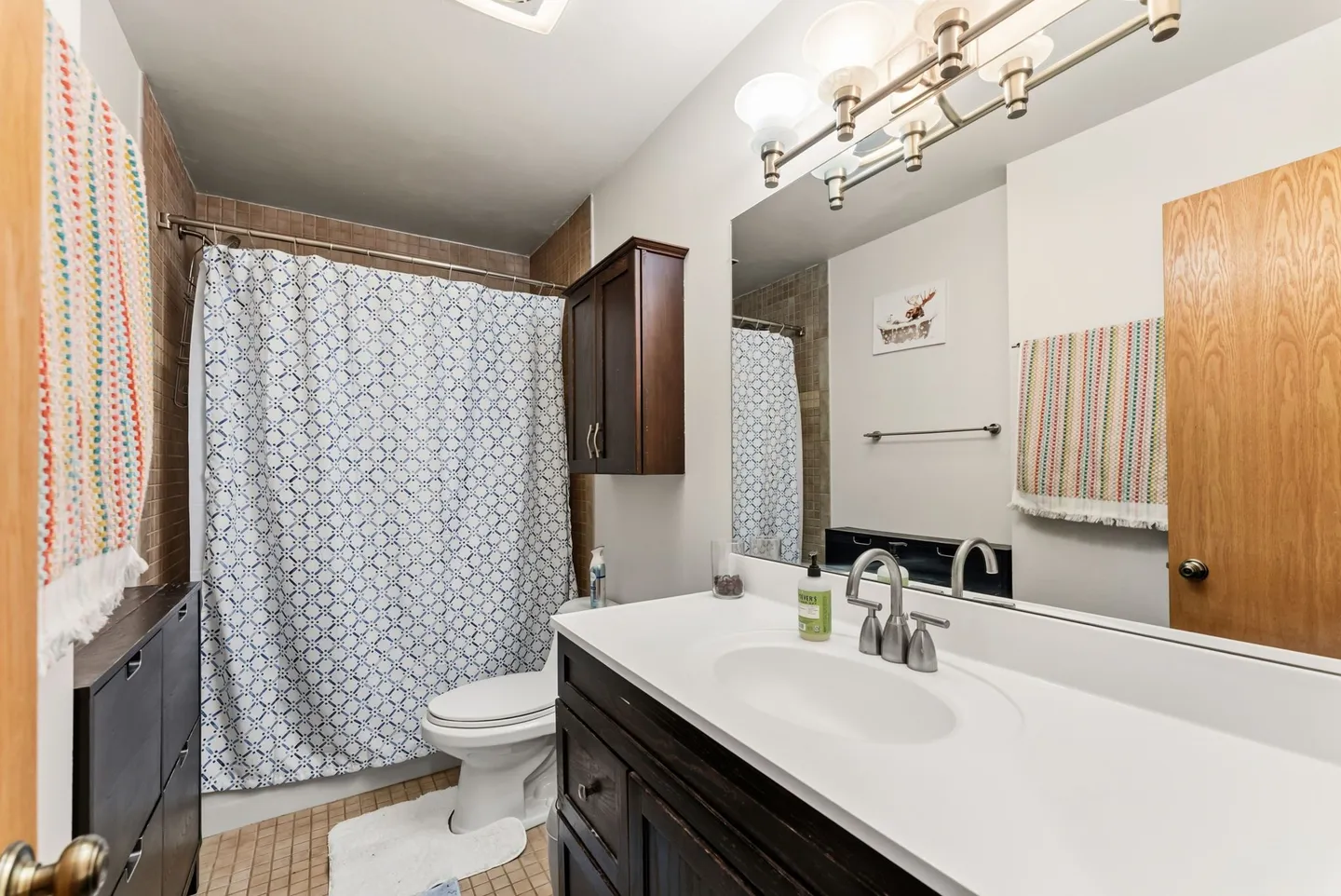 Bathroom with dark wood vanity, white countertop, large mirror, toilet, and shower with patterned curtain.